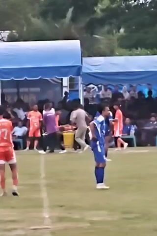 The media shows a soccer match taking place on a grassy field. Spectators are gathered behind tents, some seated and others standing. Two teams are present, one in orange jerseys with the number 77 prominently displayed on the back of one player, and another in blue and white striped jerseys with the number 19 visible on one player. 

In the background, a person appears to be injured and is being attended to by others, with a stretcher visible. This suggests a pause or interruption in the game due to the injury. The presence of tents and a large crowd indicates a community event or a local league match. The time of day appears to be afternoon or early evening, given the lighting conditions. The general atmosphere seems to be one of focused attention on the field and the ongoing events.

The scene is set in Desa Meunasah Blang, Indonesia, as indicated by the provided location context. The weather appears to be overcast or cloudy, with no direct sunlight casting harsh shadows, but the overall lighting is bright enough for the event to proceed. The visible text on the tents is in a foreign language, likely Indonesian, but it is not clear enough to read specific words or phrases. The attire of the spectators and the players suggests a warm climate.