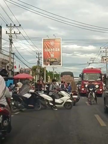 The video shows a busy street intersection during what appears to be daytime. Traffic is congested with numerous motorcycles, cars, and a large red truck. A banner hangs over the road with the word "SCORPION" and the number "16" prominently displayed. Several people are visible riding motorcycles and interacting with each other. The sky is overcast, suggesting cloudy weather. The environment seems to be urban or suburban, with buildings and utility poles lining the street. The overall impression is one of everyday activity and movement within a traffic-filled area.
