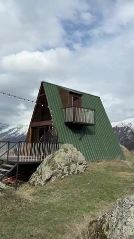 The camera slowly pans from a small A-frame house with a green metal roof to the mountains in the background. The house is situated on a rocky hill with green grass and is surrounded by large boulders. The A-frame has a small wooden balcony jutting out from the side. The mountain range behind the house is partially covered in snow. The sky is overcast with gray clouds. There is no apparent activity or people visible in the scene.