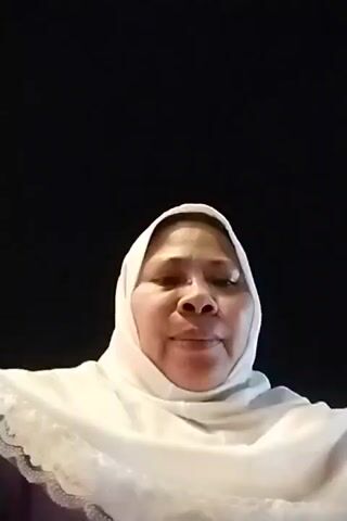A woman wearing a white hijab and a purple shirt is holding a phone and filming herself at night. She is in what appears to be an amusement park, with a brightly lit ride visible in the background. The ride is circular and has green and blue lights. The woman is smiling and looking at the camera. She is holding the phone at arm's length to record the video. The background is dark, indicating it is nighttime.