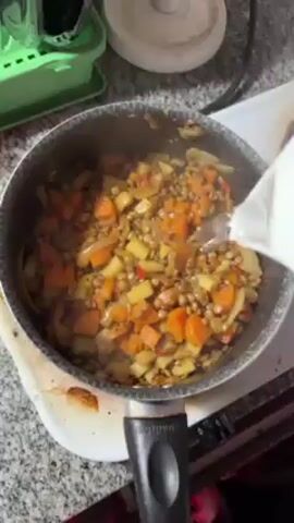 A close-up shot shows a dark gray saucepan on a white stovetop. The pan contains lentils and various diced vegetables, including carrots, potatoes, onions, and red bell peppers, simmering in a broth. Someone is pouring water into the pan from a white pitcher, creating steam. The countertop around the stovetop is speckled gray and white. A green dish rack is visible in the upper left corner, with a white object and the edge of a metal spoon visible nearby.  The stove knobs are black and located on the front. The camera angle is from directly above the saucepan.