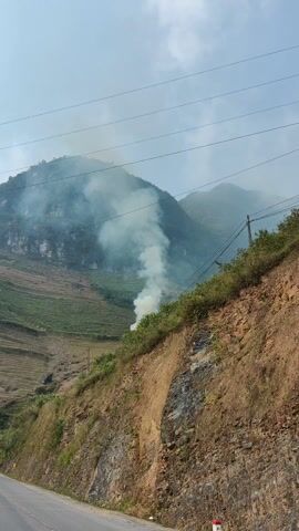 The video shows a landscape with a mountain in the background, partially obscured by fog or low clouds. A plume of white smoke rises from the hillside, indicating a fire. The hillside is terraced, with visible sections of vegetation and rocky outcrops. In the foreground, there is a road with a white guardrail, and a person can be seen walking along the road. Power lines are visible stretching across the sky. The overall impression is of a rural or mountainous area with some form of fire burning on the hillside.