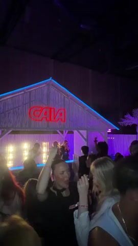 An indoor club or concert scene features a crowd dancing under purple and blue neon lighting. In the background, a stage is designed as a beach hut with a thatched-style roof and blue neon trim. A prominent red neon sign spelling "GAIA" is centered on the hut's front. On the stage, a person in a white tank top and dark shorts wears headphones, facing the crowd, possibly DJing or performing, with other individuals visible behind them. In the foreground, numerous attendees are actively dancing; a woman in a black top with pigtails smiles while raising her right arm, and a woman with long blonde hair in a white top dances beside her. The environment is dimly lit with focus on the vibrant stage lighting and the energetic crowd.