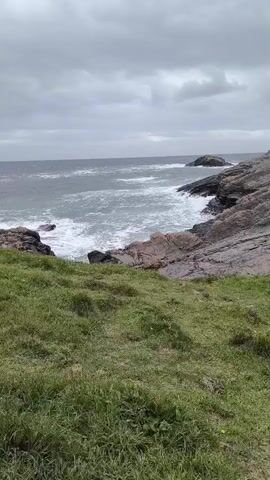 This media file depicts a rugged coastline in Tullymore, Ireland, on an overcast day. The scene is dominated by the vast, choppy ocean meeting a rocky shore. In the foreground, lush green grass covers a gently sloping hill, leading the eye down towards the water. The rocks are a mix of dark grey and reddish-brown, showing the raw power of erosion. White waves crash against the rocks, creating a dynamic contrast with the stillness of the land. In the distance, a small island or larger rock formation breaks the horizon, further emphasizing the expanse of the sea. The sky is filled with heavy, grey clouds, suggesting a cool, possibly breezy, atmosphere. There are no people or man-made objects visible, creating a sense of untouched nature and wild beauty. The overall mood is one of solitude and the untamed power of the elements. The time of day is indeterminate due to the overcast conditions, but the lighting suggests it is daytime.