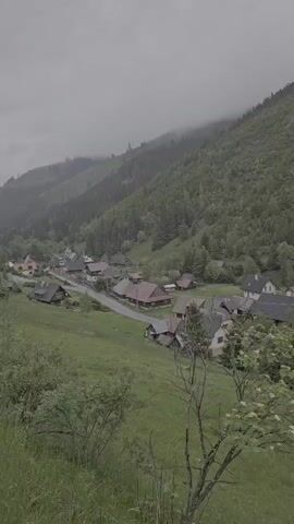 The video shows an overview of a small village nestled within a valley, surrounded by forested mountains under a cloudy sky. The village consists of various houses with different roof styles and colors, scattered along a road that winds through the valley. Lush green fields and trees surround the village, emphasizing its rural setting. In the distance, the mountains are covered in dense forests, with fog or low-hanging clouds obscuring the peaks. The overall scene is peaceful and scenic, characteristic of a remote, mountainous area.