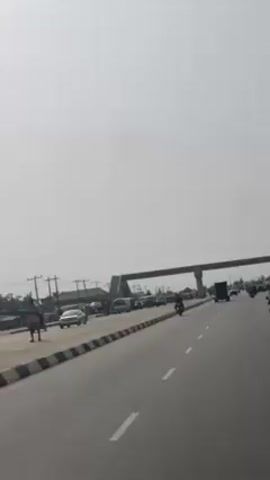 The video shows a road with traffic on both sides. On one side of the road, there are several cars and vans stopped. There are also some people standing around near the vehicles. On the other side of the road, a few cars and a motorcycle are driving. There is an overpass above the road. The sky is overcast and the horizon is hazy.