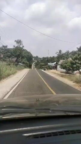 The video is a first-person perspective from inside a vehicle, looking forward. A two-lane asphalt road stretches ahead, with a yellow line dividing the lanes. The road is flanked by vegetation, including trees and bushes. On the right side of the road, there are several small houses and a parked motorcycle. The sky is overcast with clouds. The vehicle appears to be moving, as indicated by the slight blur and the perspective of the road ahead.