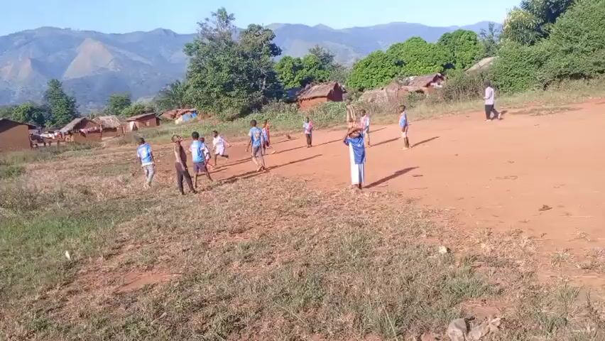 The video shows a group of children playing soccer in a village setting. There are several small, simple houses with thatched roofs in the background, along with green trees and a distant mountain range. The children are of various ages, and most are wearing colorful shirts and shorts. They are playing on a dirt field with patches of grass. In one segment, the camera focuses on the ground where a soccer ball and some trash are visible. A child then walks past this area.