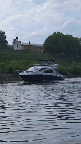 A large, two-level, white and blue yacht is seen moving across the rippling surface of a river. The vessel is propelled forward with a slight wake trailing behind. On the top deck, figures are vaguely visible under the shaded awning. A small Russian flag is attached to the stern. The riverbank features a grassy hill, punctuated by trees, bushes and a large brick building adjacent to a smaller, white, domed structure that appears to be a church or chapel. The sky above is overcast.