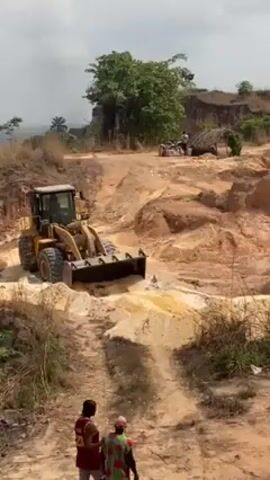 The video depicts a yellow front-end loader working in a dusty, sandy quarry or construction site. The loader is moving on an uneven, unpaved path. In the foreground, two people, one wearing a red shirt and the other wearing a brightly colored dress, stand and appear to be watching the loader. In the background, there is a makeshift shelter and some supplies. The landscape includes trees, hills, and a hazy sky. The overall impression is that of an active work site in a rural area.