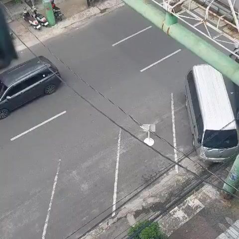 This video captures a street scene in Pematang Siantar, Indonesia. The primary focus is on a small, brightly colored van with a black roof, decorated with various decals and imagery, moving down a paved road. The van appears to be a form of public transportation, possibly a minitrans or a similar local vehicle. It has its side door open, suggesting it might be picking up or dropping off passengers.

In the background, a large silver van is parked on the side of the road. Further down the street, a blue truck is visible, as well as a smaller motorized vehicle that resembles a scooter or a small utility cart. A pedestrian walkway runs alongside the road, with some foliage and wires overhead. A green bridge or overpass structure is prominent in the upper right portion of the frame.

The activity appears to be normal daytime street traffic. The lighting suggests it is daytime, with no obvious indications of specific weather conditions beyond it being a typical urban environment. The scene is likely taking place in a commercial or residential area of the city, given the presence of different types of vehicles and overhead wires commonly found in urban settings. No text is clearly legible on the vehicles or surroundings within this clip.