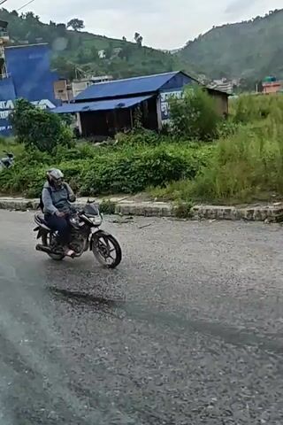 A motorcyclist wearing a helmet rides a motorcycle on a wet road. The rider is leaning to their left as they navigate a curve. Buildings and lush green vegetation are visible on the hillside in the background. The sky is overcast. The video appears to be filmed from inside a vehicle, with a curtain visible in the foreground.