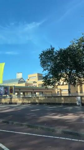 This video captures a sunny day in The Hague, Netherlands, likely mid-morning or early afternoon given the bright sunlight and clear blue sky with wispy clouds. The scene features the Kunstmuseum Den Haag, a prominent art museum, with its distinctive yellow brick facade and modern architectural elements.

Several flags are prominently displayed outside the museum. A large, red vertical banner reads "KUNST MUSEUM" and "DEN HAAG". To its right, a yellow banner also displays "MUSEUM", and a light green banner shows "MUSEUM" as well. In front of the museum, three large posters advertise exhibitions, one featuring colorful abstract art, another with a silhouette of musical instruments and text, and a third with a portrait and vibrant colors.

The area appears to be a public space with a paved walkway and street. A few people are visible on scooters, indicating casual transportation and movement around the city. Bicycles are parked at racks near the museum entrance, suggesting a common mode of transport for visitors. Red and white striped barriers are also visible, potentially indicating construction or a temporary event. The overall atmosphere is calm and pleasant, with the bright weather contributing to a cheerful mood. The architecture of the museum itself is a key visual element, with its clean lines and warm-toned brickwork standing out against the blue sky.