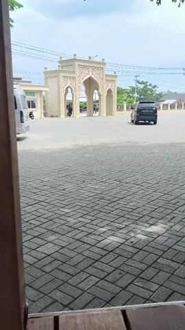 A black SUV drives through a paved courtyard in front of a mosque with a large arched entrance and a dome. A white minivan is parked to the right of the frame. The sky is overcast.
