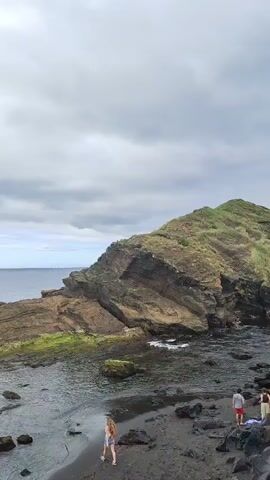 This media file captures a scenic coastal view with a dramatic cliff face and the ocean under a cloudy sky. In the foreground, a person is walking on a dark, rocky beach with shallow water. Further out in the ocean, two boats are visible. The first boat is a small inflatable raft with several people on board, seemingly enjoying a leisurely ride. The second boat is a sailboat with its sails up, moving gracefully across the water. In the background, a lush green hillside slopes down towards the sea, with a distant town or village nestled along the coastline. The overall atmosphere is peaceful and serene, with the overcast sky adding a touch of dramatic beauty to the landscape. The scene suggests a day of exploration and enjoyment of the natural surroundings.