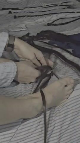 The video shows a person's hands and feet on a bed covered with a striped comforter. The person's hands are braiding long, dark hair extensions. Multiple hair extensions are laid out on the bed, some with beads. One of the person's feet is visible with a hair extension wrapped around the ankle. The overall scene suggests a person is either creating or preparing hair extensions.