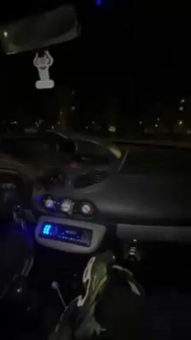 The video is shot from inside a parked car at night. The interior of the car is dimly lit with a bright blue light emanating from the radio panel. A person is seen sitting in the driver's seat. The camera pans from the radio up towards the windshield showing the street outside and apartment buildings lit by streetlights. A hanging air freshener is visible from the rearview mirror. The camera comes back down toward the dashboard. Loud music is playing throughout the video.