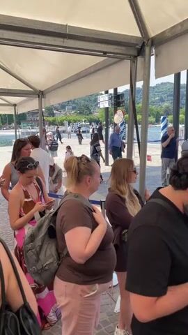 The video shows a crowd of people waiting in line at a waterfront location. Most individuals are standing under a white canopy, using their phones or looking around. The environment includes glimpses of the lake, buildings, and other people passing by. The line appears to be leading to an entrance, as indicated by a sign.