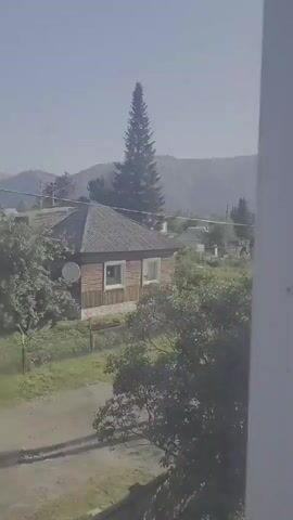 The video shows a rural landscape with several houses and trees. The sky is clear and blue, with mountains in the distance. The houses appear to be made of wood and have pitched roofs. There is a tall, thin evergreen tree in the foreground, and other trees and bushes are scattered throughout the scene. A dirt road runs through the middle of the frame, and a fence can be seen along the side of the road. The overall impression is one of a peaceful, rural setting.