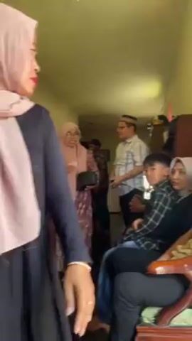 The video shows the interior of a home in Indonesia, with several people present. Some are standing in a hallway, while others are seated in the living room. Most of the women are wearing hijabs, and some of the men are wearing traditional Muslim caps. A woman in a pink patterned dress is handing money to a boy who is sitting on a woman's lap. There is furniture and other household items visible in the background. The walls are painted yellow. The atmosphere appears to be somewhat formal or celebratory.