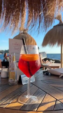 A close-up shot focuses on a red cocktail with an orange slice and a black straw, resting on a wooden table. The glass is beaded with condensation. To the left, a bottle of sparkling water and salt and pepper shakers are visible. A menu card with "CHIRINGO" and "SWEET COCKTAILS" printed on it stands behind the drink. In the background, out of focus, are people sitting at tables, a beach with thatched umbrellas, and the ocean under a blue sky. The lighting suggests it is daytime.