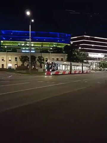 The video shows a tram at night. The tram is white with red stripes and has its interior lights on. It is on a street with multiple lanes. In the background, there is a building with blue and green lights. There are streetlights and other buildings visible. The sky is dark. The tram appears to be moving.