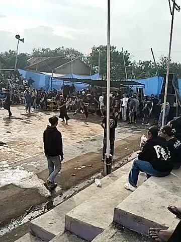 A group of people are gathered in an outdoor court. In the center of the court, two individuals are engaged in what appears to be a performance or a fight, with one person in a black outfit and the other in a dark outfit. Surrounding the court are spectators seated on bleachers or standing behind a blue tarp. The court itself is a concrete surface with some debris and puddles of water. Two tall poles with lights and speakers are positioned on either side of the court. The sky is overcast.