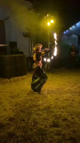 A woman wearing a floral headdress, black crop top, and flowing green skirt is performing with fire. She is spinning what appear to be fire poi, creating large circles of flame around her. She is outdoors at night, on a grassy area in front of a stage setup. There are lights illuminating the stage and the performer. In the background, there is a building decorated with string lights. Several people are seated or standing in the background, watching the performance. The overall scene suggests an outdoor event or festival.