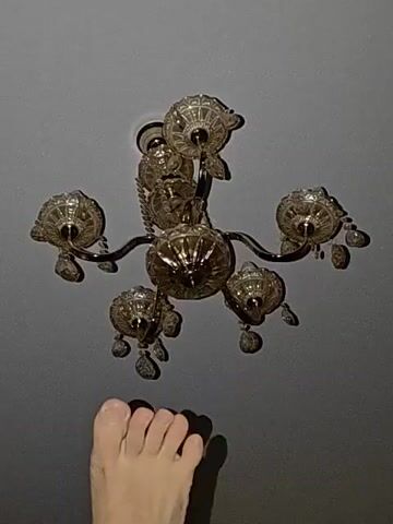 A foot is positioned in the lower left corner of the frame, facing right. Above the foot and occupying the majority of the frame is a chandelier hanging from a gray ceiling. The chandelier is ornate, with multiple arms, each holding a decorative glass shade. Crystal-like embellishments hang from the bottom of the chandelier. The lighting in the scene is dim.