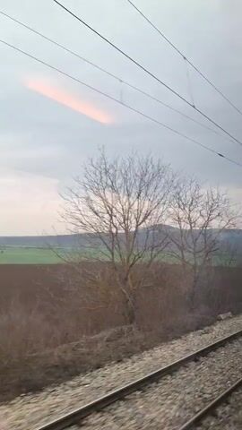 The video shows a view from inside a moving train, looking out of the window. Outside, the landscape consists of railway tracks, a low hedge, a bare tree, fields, and a distant hill under a cloudy sky with a hint of sunset. The overhead electrical wires for the train are also visible. The camera then pans upward to show the interior of the train carriage, which is mostly empty, with blue seating and an overhead compartment.