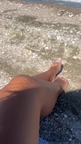The media file displays a close-up view of a person's legs and feet, with manicured toenails, resting on a pebble beach. The scene transitions to a broader view of the beach and the sea. The water is calm with gentle waves, and the sky is a clear blue, suggesting a sunny day. The beach is covered in a multitude of small shells and pebbles. In the distance, a person can be seen on a paddleboard or a small boat. The overall mood is relaxed and peaceful, evoking a sense of a pleasant day at the seaside. There is no visible text in the media file.