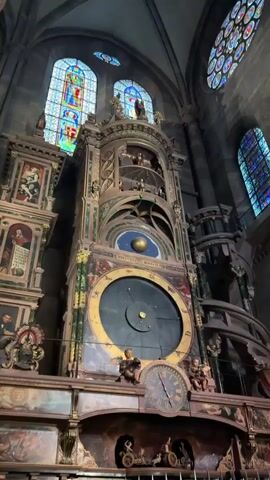 The video shows a close-up view of an ornate astronomical clock inside a grand building, likely a cathedral or church. The clock is very large and intricate, with multiple dials and moving parts. The main dial is a deep black with a golden outer ring displaying astrological symbols and Roman numerals. A golden sphere is positioned above this main dial, set against a starry blue background, possibly representing the celestial sphere. Smaller dials and figures are visible on the clock's structure, including what appear to be cherubic figures and depictions of historical scenes. Stained-glass windows, illuminated by natural light, are visible in the background, adding to the architectural grandeur of the space. The camera slowly zooms in and pans slightly, highlighting the detail and craftsmanship of the clock. The overall impression is one of historical significance and mechanical complexity.