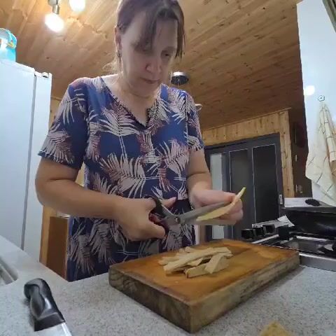 A woman stands in a kitchen, wearing a blue and white patterned dress, and uses scissors to cut strips of food on a wooden cutting board. The kitchen has wooden paneled walls and ceiling, a white refrigerator, a stove with a black pan on it, and a white countertop.