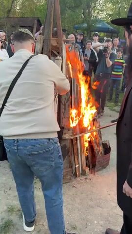 In a medium-shot outdoor scene, a group of people, many wearing kippahs or traditional Jewish hats, gather around a bonfire made of wood scraps. The bonfire burns intensely, with orange and yellow flames reaching up the structure. Several men in the foreground hold long, thin sticks with flames at the end, adding them to the larger fire. One man is dressed in a dark suit and hat, and another wears a tan sweater and jeans. The crowd behind them includes people of various ages and genders, some recording the event with their phones. The background features trees, a building, and a blue sky, suggesting it's late afternoon or early evening. The ground is dirt with some scattered debris. The atmosphere seems celebratory or ceremonial.