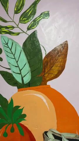 The video shows a close-up, slightly shaky, horizontal pan of a painting on a light purple wall. The painting depicts a still life of a terracotta-colored vase with a bright orange rim, filled with various green and brown leaves. In front of the vase, to the left, is a round, orange object that appears to be a fruit, possibly a tomato, with green leaves on top. To the right of the vase, a beige object with a patterned strap is partially visible, suggesting it is a bag or basket. The painting style is bold with thick brushstrokes, and the lighting seems to be natural, casting soft shadows. A hand holding a painting tool, possibly a brush or palette knife, briefly enters the frame from the left at the 00:02 mark, suggesting someone is in the process of painting or adding details. The painting itself is colorful and stylized, with a focus on organic shapes and vibrant hues.
