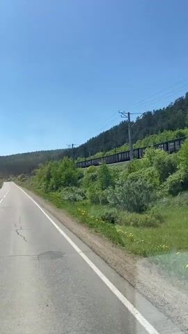 The video shows a view from a moving vehicle on a paved road. The road is cracked and has a white line dividing the lanes. On the right side of the road, there is a grassy embankment with yellow flowers and a line of trees. Further in the distance, a train with open cars is visible on a railway track, with power lines running overhead. The sky is clear and blue. On the left side of the road, there is a body of water, possibly a river, bordered by a metal guardrail and trees. The vehicle continues to move, and the scenery shifts slightly. The interior of the vehicle is briefly visible in the foreground, showing part of a pillar and window frame. The video then focuses on the water and trees on the left side as the vehicle moves past. The overall environment appears to be rural and natural, with abundant greenery and clear weather.