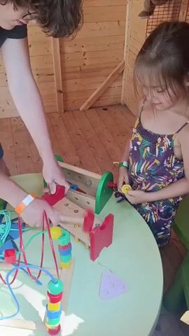 A young girl with dark hair, wearing a colorful floral dress, is playing at a light green circular table with a wooden toy. The toy has a green and red frame, a wooden cylinder in the middle with holes, and several colorful pegs. The girl is using her hands to manipulate the pegs in the toy. To her left, a person with dark hair, wearing a black t-shirt and blue shorts, is also interacting with the toy, placing their hands near the pegs. On the table, there are other children's toys, including a wire bead maze with colorful beads, some blue and red geometric shapes, and a pink triangular shape with three holes. The background appears to be a room with light brown wooden walls and floor, possibly a playhouse or a cabin. The lighting is bright and natural.