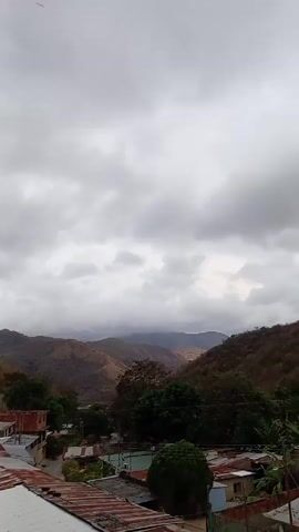 The video shows a cloudy sky over a valley. There are mountains in the distance, covered in sparse vegetation. In the foreground, there are some rooftops and trees. The sky is filled with grey clouds, obscuring most of the sunlight. The video is static.