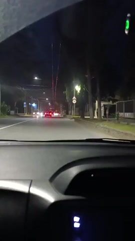 The video shows a first-person view from inside a car at night. The car is driving on a wet, paved road lined with trees and streetlights. There are other cars on the road, some with their headlights and taillights illuminated. A speed limit sign is visible on the right side of the road. A person can be seen standing on the sidewalk on the right. The dashboard of the car is visible in the foreground.