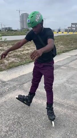 A young Black male wearing a green helmet, black t-shirt, purple pants, and inline skates is standing on a paved surface. He has a black bag slung across his chest. He is balancing on one skate, with his arms outstretched. The background shows a cloudy sky, some grass, a body of water, and buildings under construction.