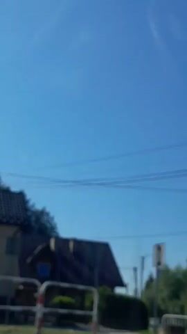 A blurred video captures a daytime outdoor scene. A blue sky is visible. In the foreground, a metal barrier with red and white markings and a circular sign with some text is present. Behind the barrier, a green bench can be seen. Further in the background, a house with a dark, sloped roof and beige walls is visible. Trees and other buildings can also be seen in the distance. The video is shaky, indicating it was likely filmed from a moving vehicle.