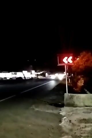The video captures a nighttime view of a road. A traffic sign with three red arrows pointing left is visible on the right side of the road. The arrows are lit, indicating a sharp left turn ahead. Cars are driving on the road, their headlights illuminating the scene. The environment appears to be semi-urban or rural, with some buildings and trees visible in the background. The road surface is asphalt, and there is a sidewalk or shoulder on the right side of the road where the camera is positioned. The overall impression is one of navigating a road at night, with clear directional guidance provided by the illuminated sign.