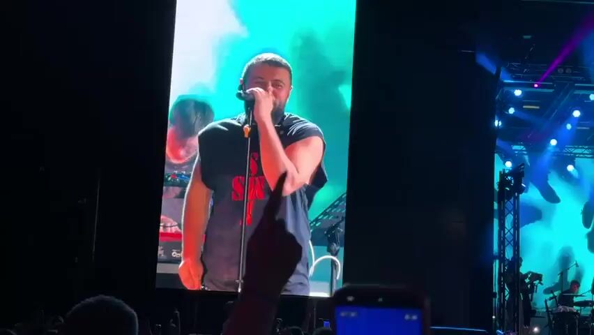 A concert is taking place on a brightly lit stage. The stage has a large screen displaying a close-up of a man singing into a microphone. The man is wearing a black sleeveless shirt with red lettering on it. In front of the screen, a band is performing. There are at least three people on stage, playing instruments that appear to be guitars and a keyboard. The stage is illuminated by numerous blue and purple lights, and there is smoke or fog on stage. Below the stage, a large crowd of people are watching the performance, many of them holding up their phones to record. The audience is densely packed, and the overall atmosphere appears to be energetic.