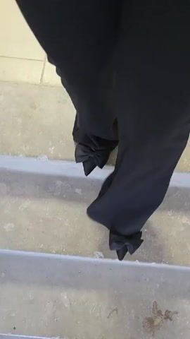 The video shows a close-up view of a person's legs from the knees down, wearing black pants and black shoes with bows on the heels. The person is walking down a set of concrete stairs. The lighting is dim, and the background is out of focus. The camera angle is from above, looking down at the person's legs and feet. The person's movements are fluid and rhythmic, indicating they are walking with ease. The stairs appear to be indoors, possibly in a building or a home. There is some debris on the stairs, but it is not clearly identifiable.