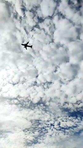 The video shows a plane flying through a cloudy sky. As the camera pans down, buildings and trees come into view. At the bottom of the frame, a person is seen standing outside. The sky is filled with dense clouds, and the sun is partially obscured.
