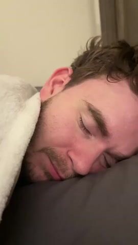 The video shows a close-up of a man sleeping on his side on a dark pillow. He is partially covered by a white fuzzy blanket. His eyes are closed, and he has a light stubble beard. In the last few frames, a hand enters the frame from the top and gently pokes the man's forehead, causing him to smile and his eyes to squint shut.