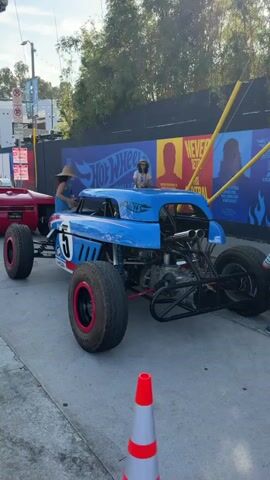 A person wearing a straw hat and a blue tank top stands on the right side of a blue and white hot rod car. The car has the number 5 on its side and large, chunky tires with red rims. The car has an open engine compartment, exposing the engine. In the background, there is a red car that is more rounded in shape, also with large tires. Behind the cars, there is a black wall with large "Hot Wheels" logos and posters. The environment appears to be outdoors, possibly a street or event space, with some trees and lighting visible. A traffic cone is in the foreground of the shot.