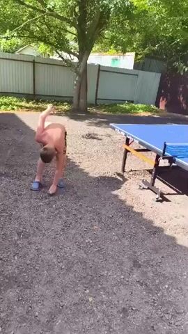 A young boy, shirtless and wearing patterned shorts, plays table tennis outdoors. He is in the process of hitting the ball with a paddle. Another person is partially visible in the background, also near a table tennis table. The scene is set in a yard with gravel on the ground, trees providing shade, and fences in the background. The tables and paddles are standard equipment for table tennis.