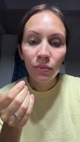 A fair-skinned woman with light-brown hair in a pale yellow t-shirt is seen in a medium close-up shot, facing the camera, in a bathroom setting. She is wearing a silver ring and a pink smart watch. Her face appears to have a white cream or lotion applied to it. She rubs her hands together and then presses them to her face, covering her eyes and mouth with her hands. A cabinet or wall is visible in the background. The lighting is bright.