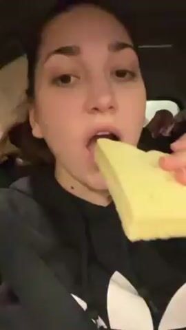 A woman with brown hair and no visible makeup is in a car. She is wearing a black hoodie with a white logo. She takes a bite out of a large, rectangular, layered piece of what appears to be pastry or cake. She chews, her eyes widen, and her expression conveys surprise or shock. The background shows parts of the car interior.